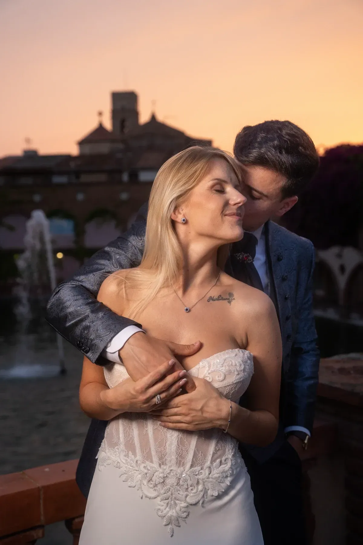 Novios se abrazan al atardecer, cerca de una fuente. El hombre besa a la mujer en la mejilla. Edificio al fondo.