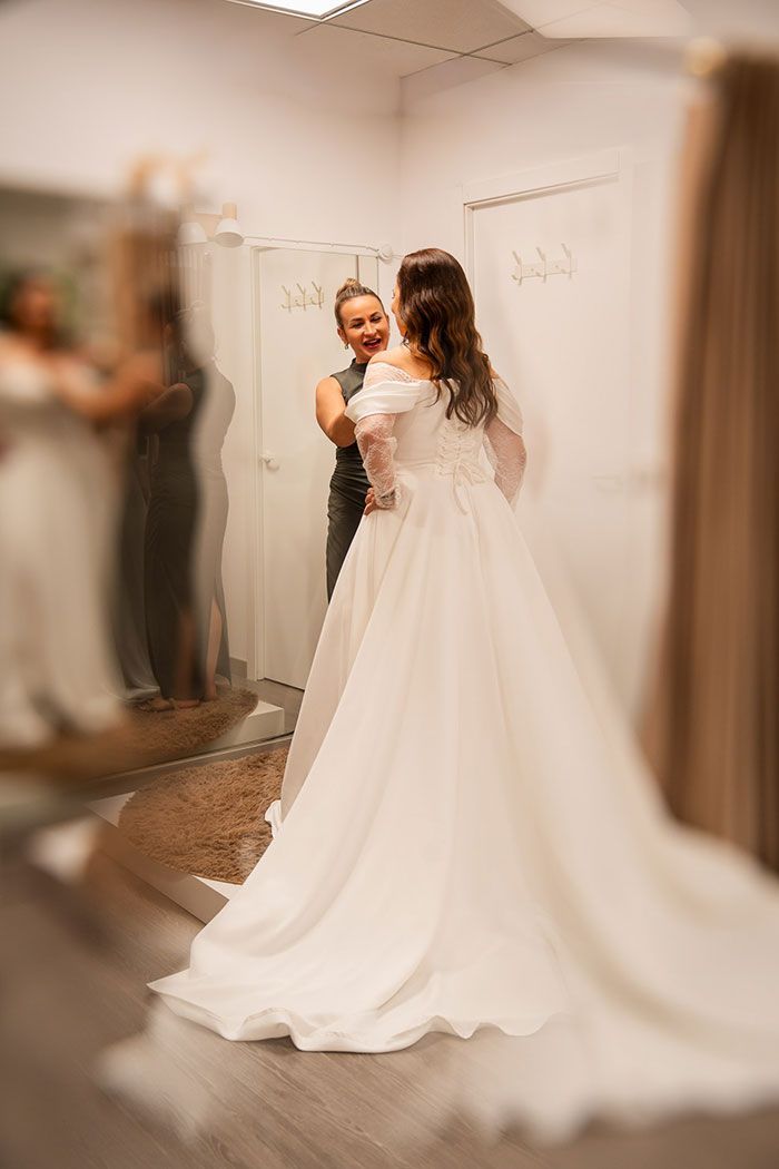 Mujer con vestido de novia en una tienda de novias, mirándose en el espejo con un asistente, sonriendo.