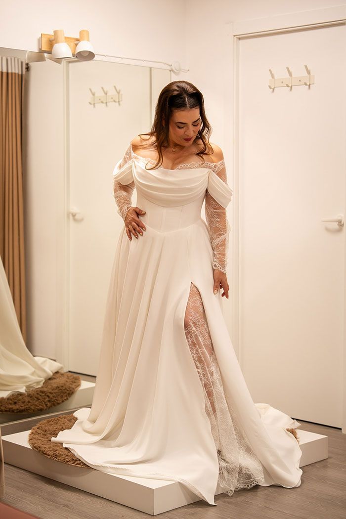 Mujer probándose un vestido de novia blanco con mangas de encaje y una abertura hasta el muslo, en un probador.