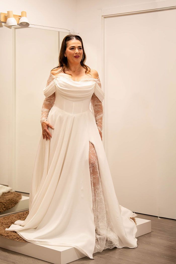 Mujer con un vestido de novia blanco con mangas de hombros descubiertos y una abertura hasta el muslo, de pie en una tienda de novias.