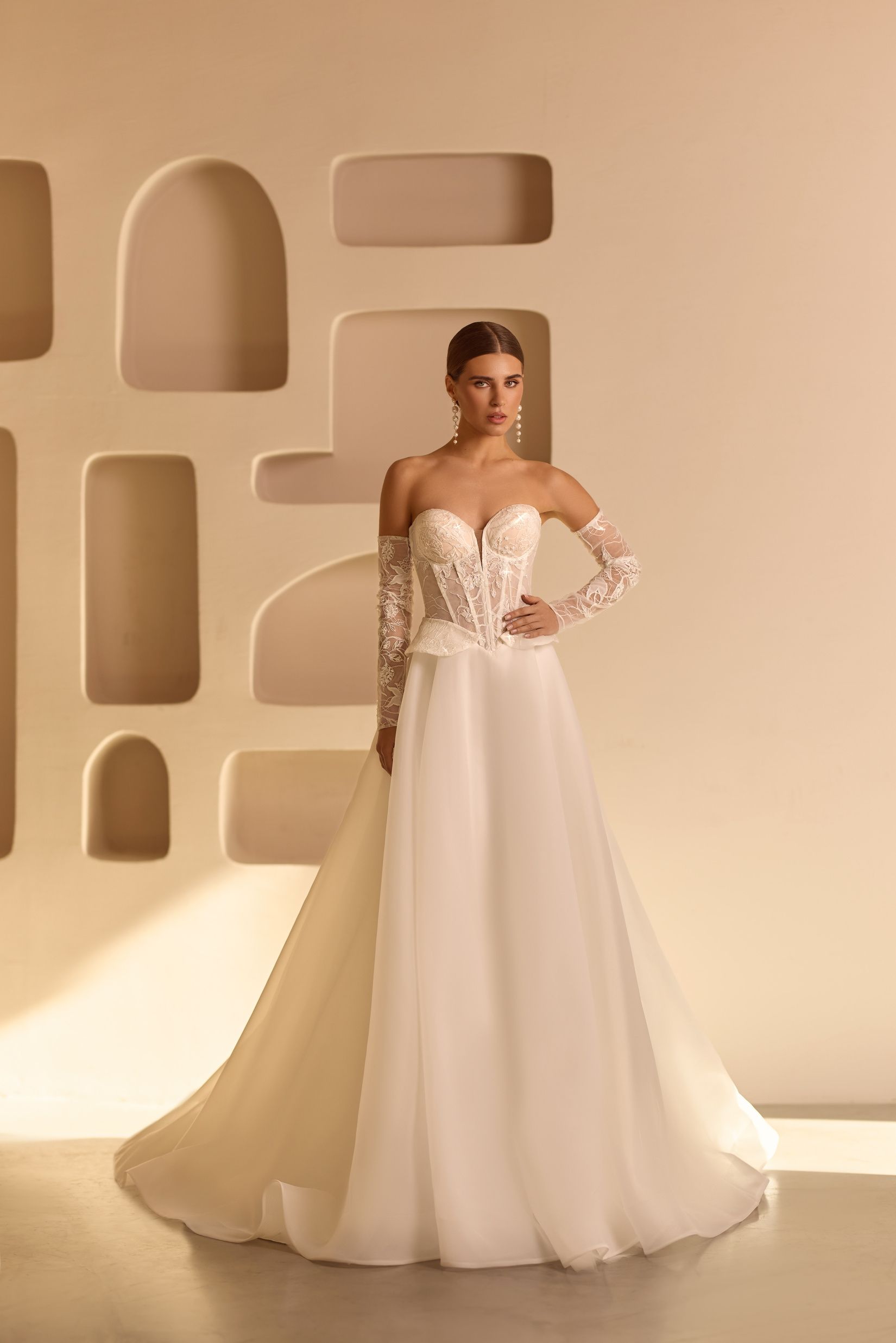 Mujer con vestido de novia sin tirantes con mangas de encaje, de pie frente a una pared beige con recortes arquitectónicos.