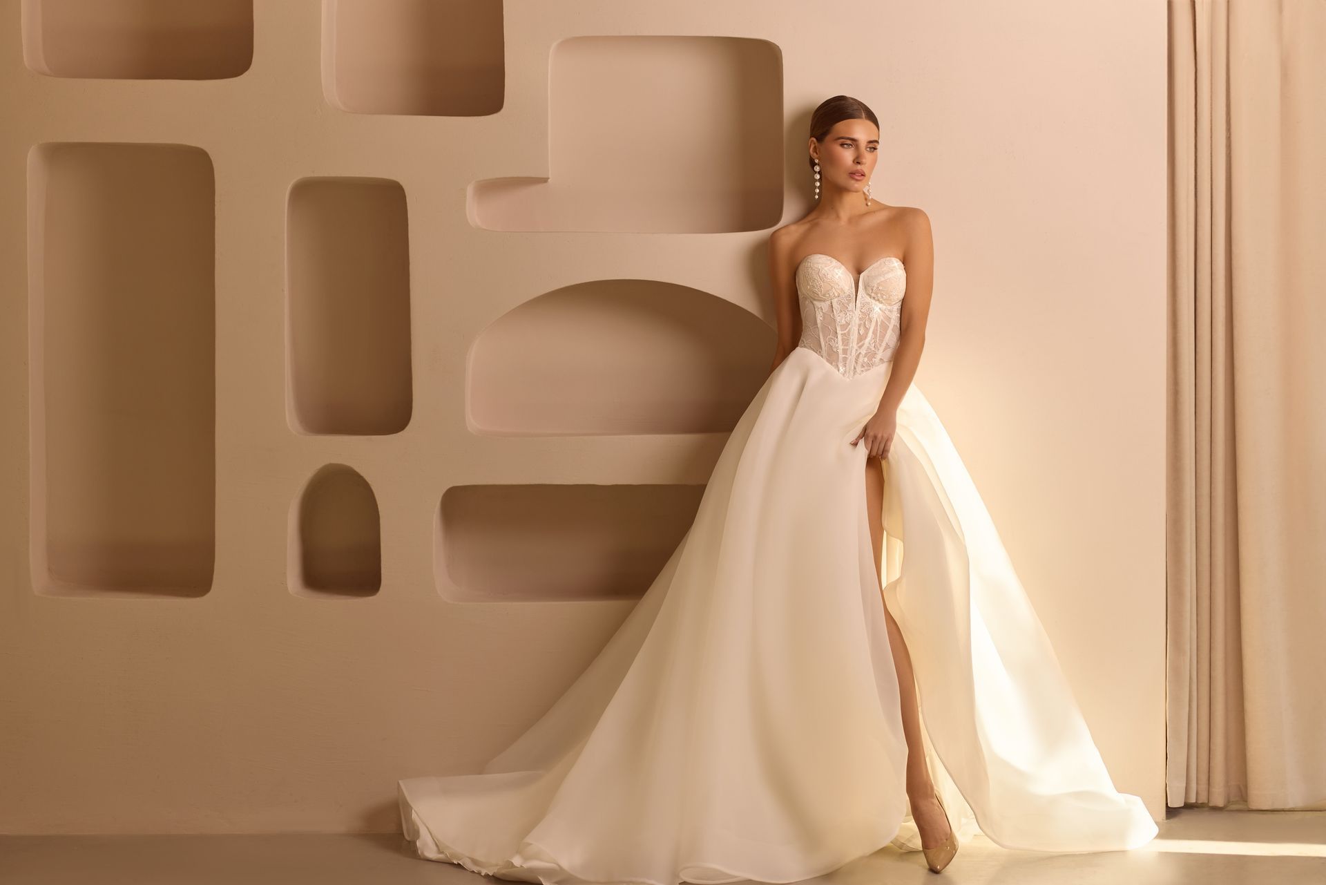 Mujer con un vestido de novia blanco sin tirantes y con abertura alta, de pie junto a una pared decorativa.