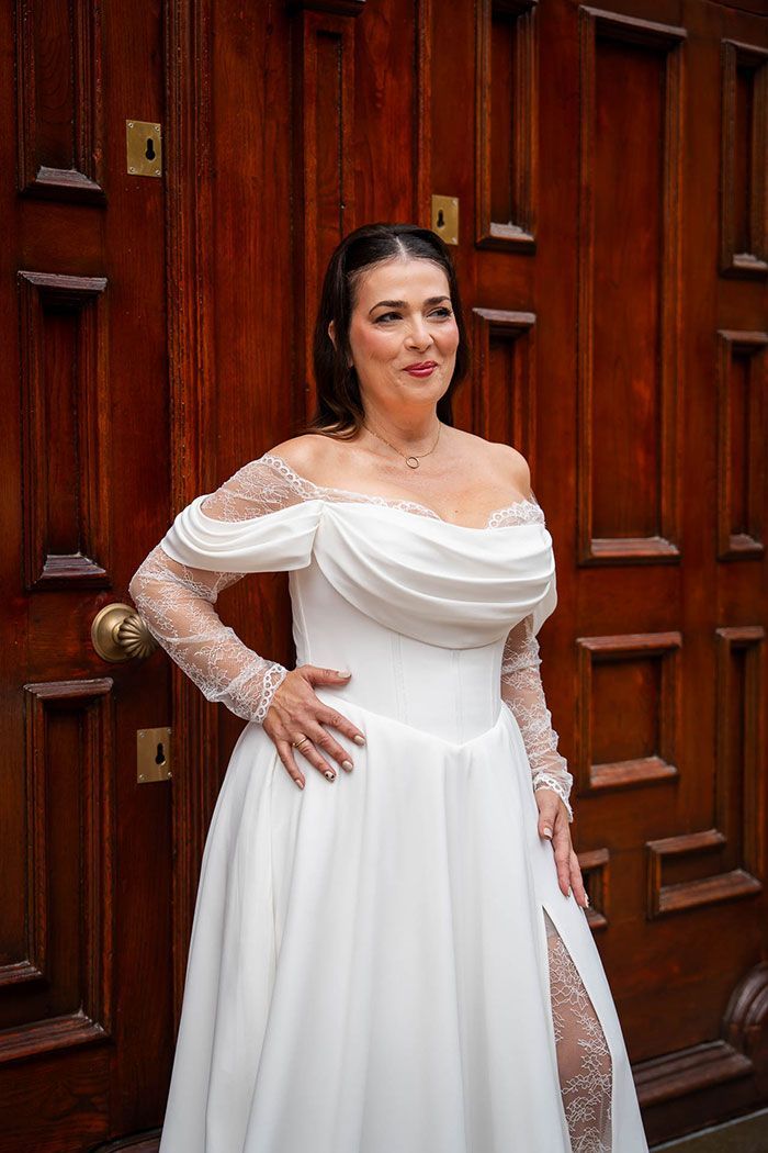 Una mujer con un vestido de novia blanco está de pie cerca de una puerta de madera, con la mano en la cadera y sonriendo.