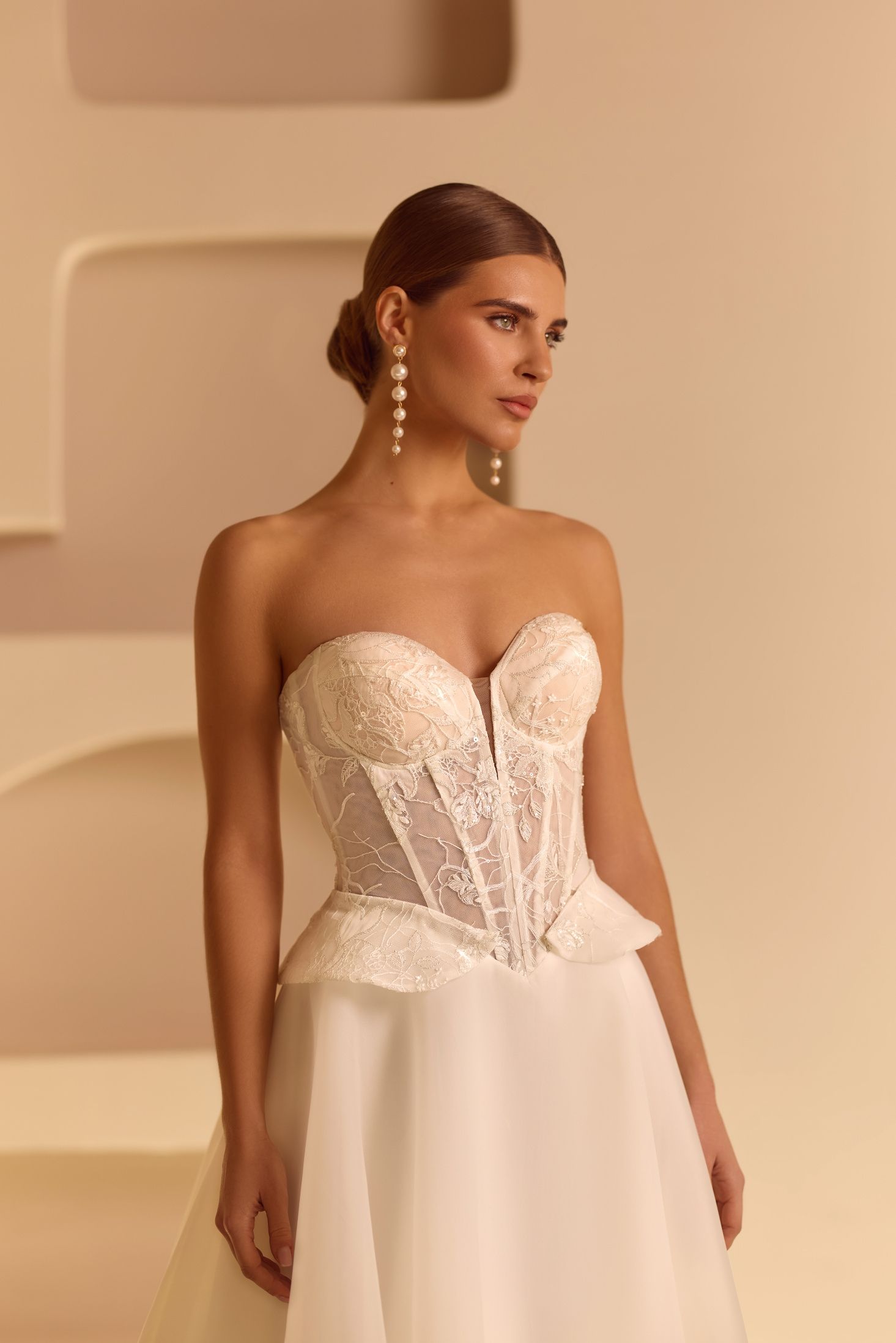 Mujer con vestido de novia blanco sin tirantes, peinado recogido, pendientes de perlas, fondo beige.
