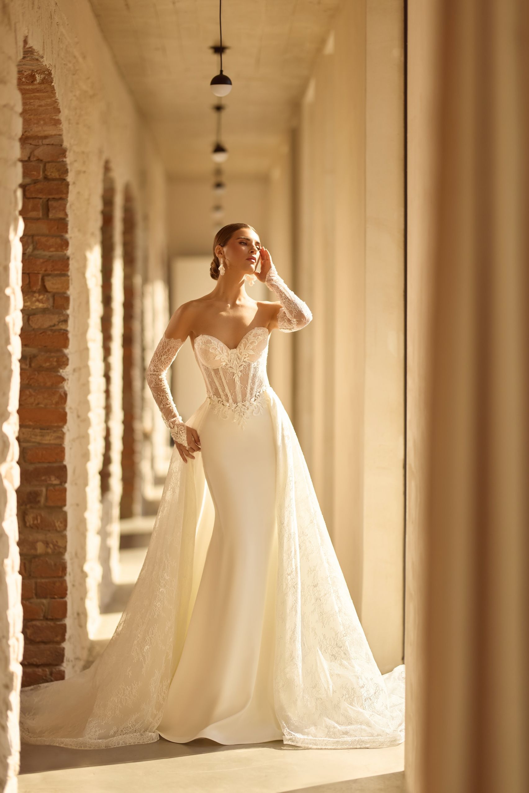 Mujer con un vestido de novia blanco con cola larga, de pie en un pasillo iluminado por el sol con arcos de ladrillo.