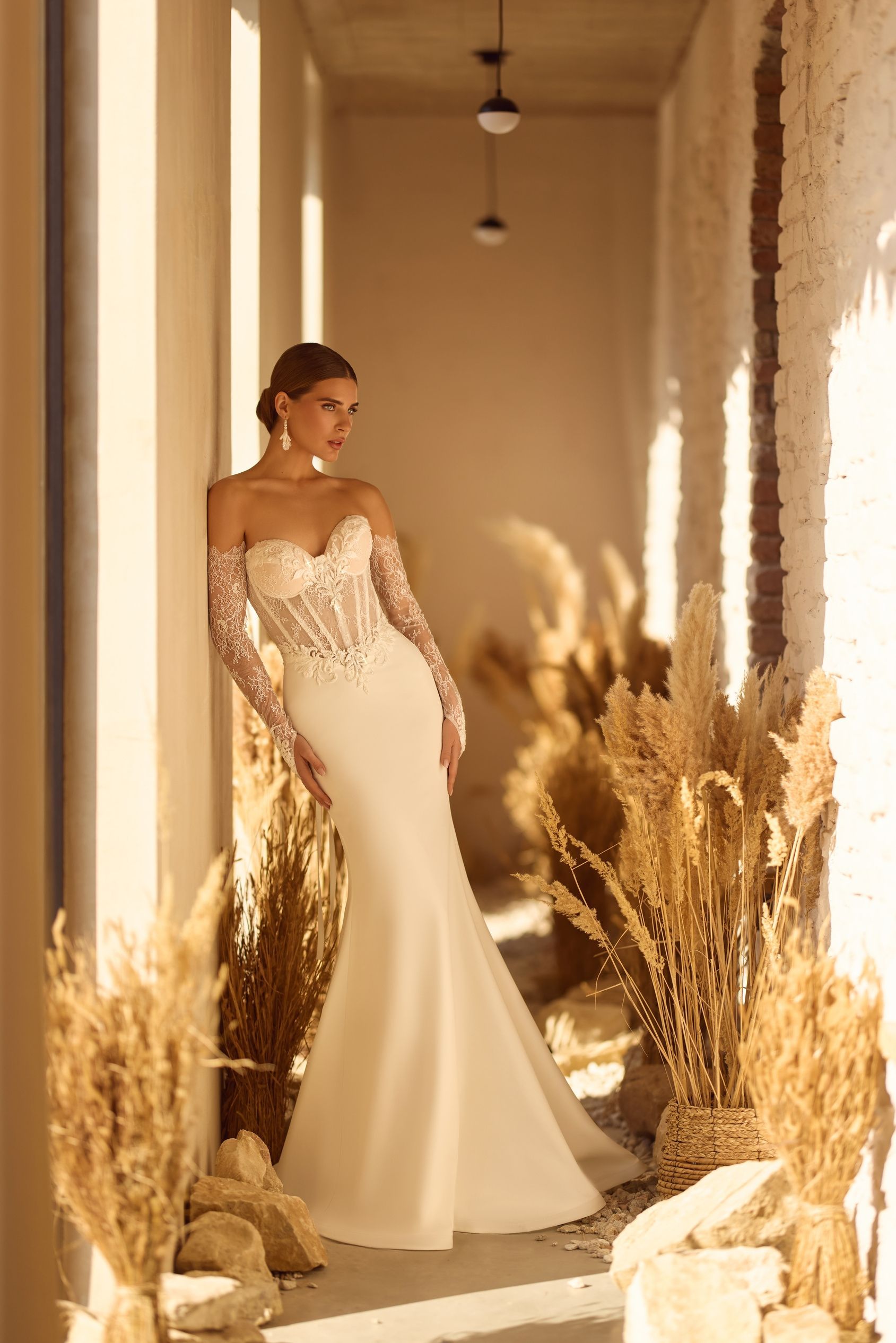 Mujer con un vestido de novia blanco con mangas largas, apoyada contra una pared, junto a plantas secas.