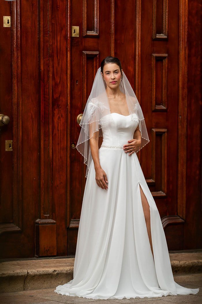 Novia con vestido de novia blanco con abertura, de pie frente a una puerta de madera. Lleva velo.