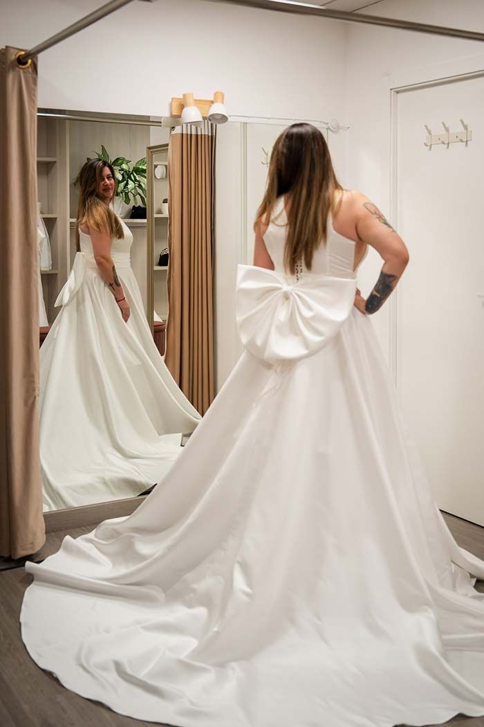 Mujer con vestido de novia con lazo grande, probándoselo en una tienda, mirándose en el espejo.