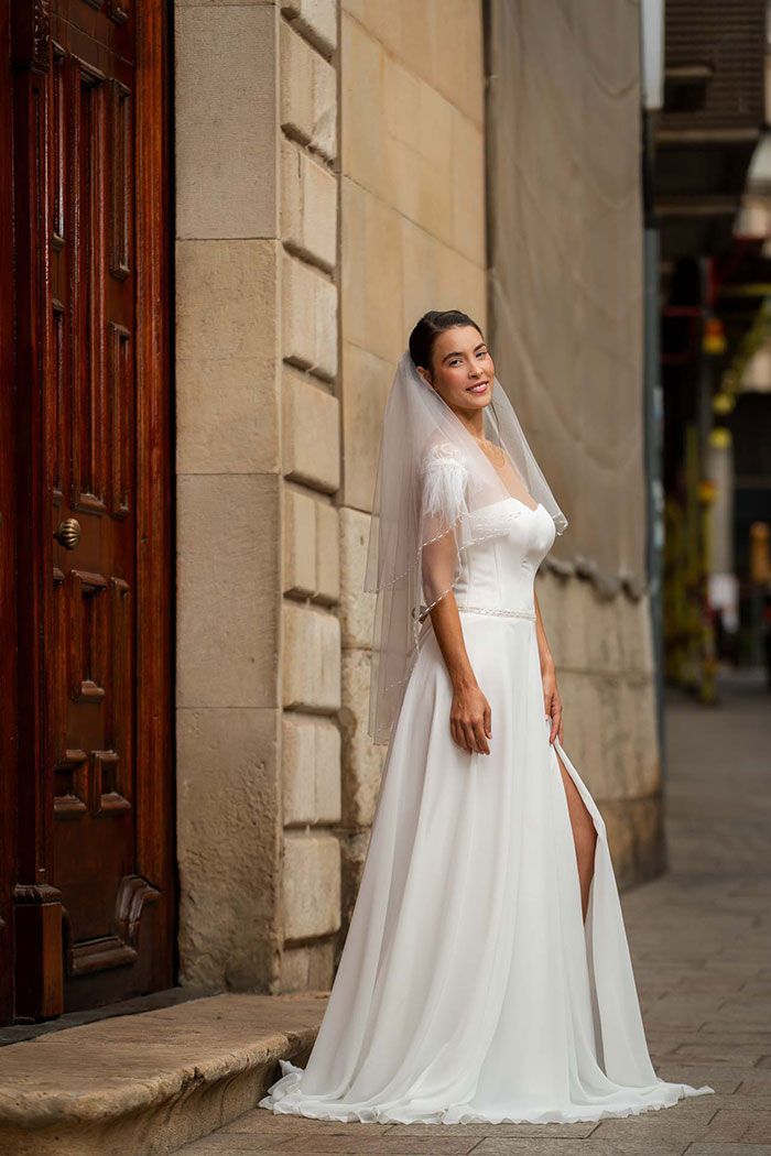 Novia con vestido de novia blanco y velo, de pie junto a un edificio.