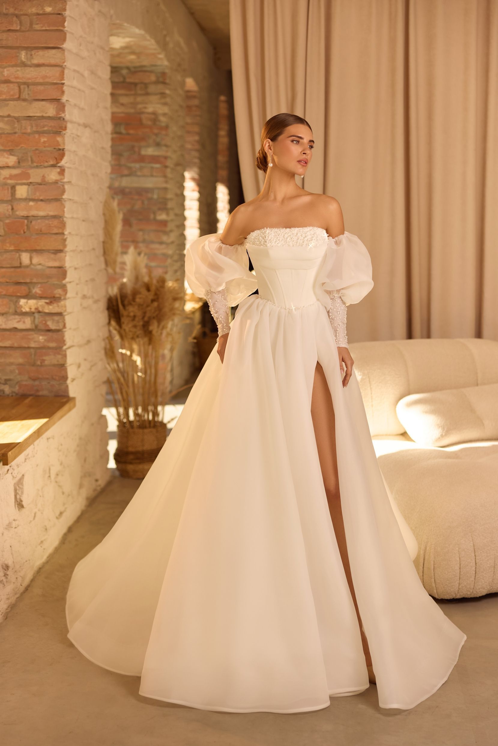 Mujer con un vestido blanco con hombros descubiertos y abertura alta, de pie en el interior cerca de una ventana y un sofá.