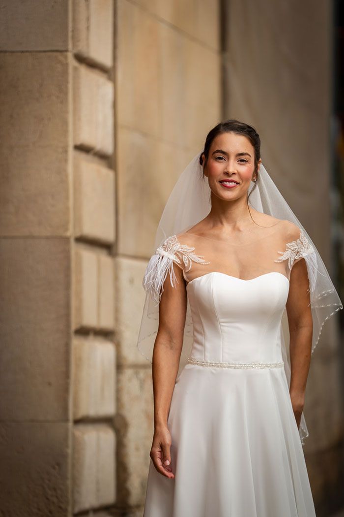 Novia con vestido blanco con mangas de encaje y velo sonríe frente a un muro de piedra.