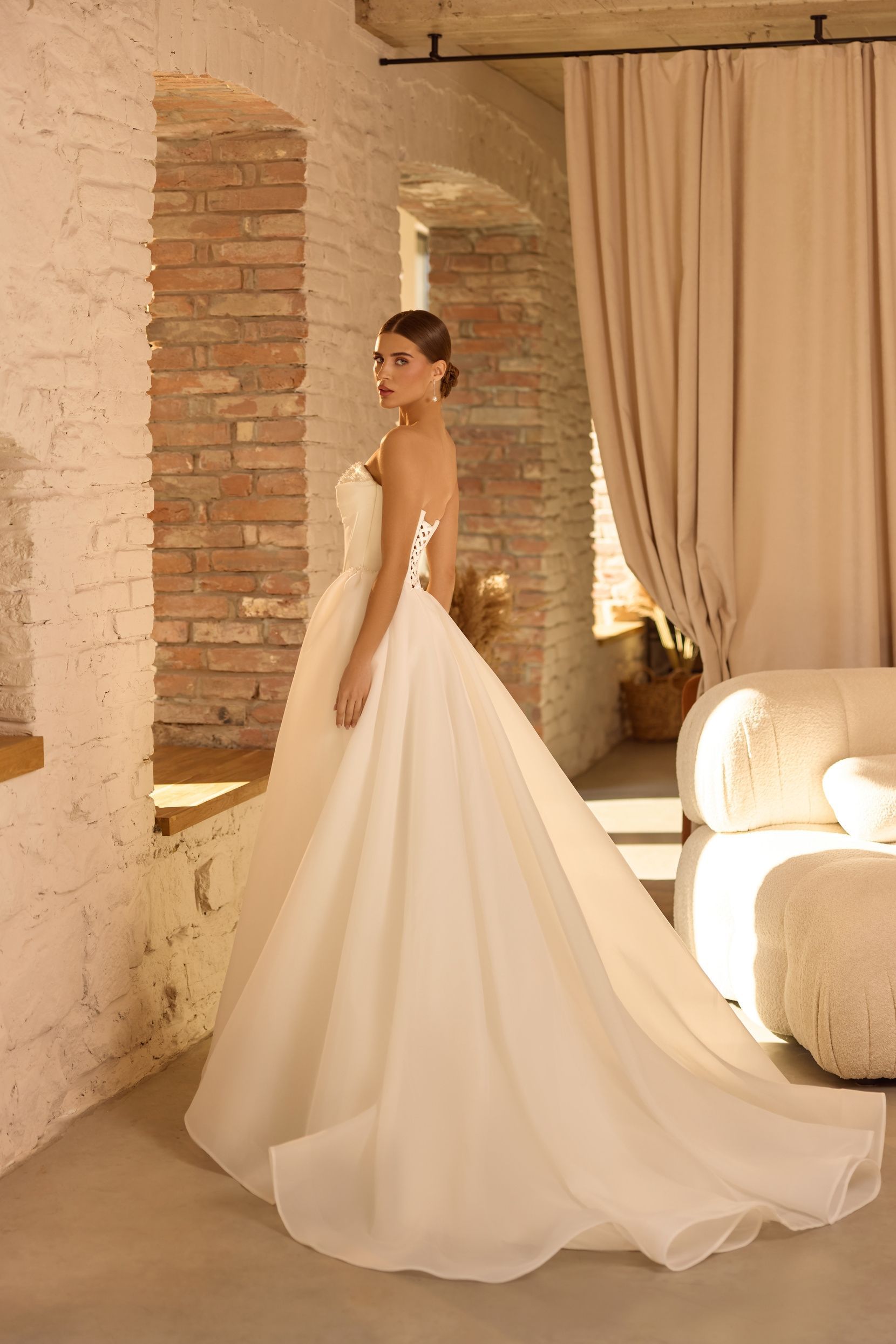 Mujer con vestido de novia sin tirantes con cola de pie cerca de una pared de ladrillos y una ventana, interior elegante.