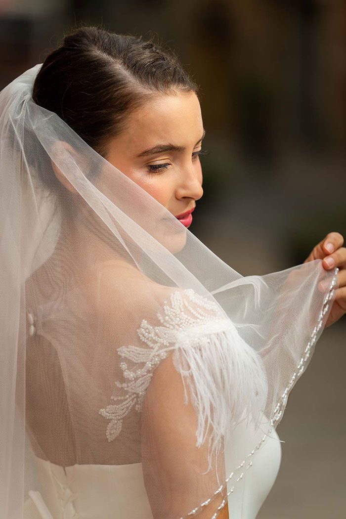 Novia con vestido blanco, velo en la mano y mirada baja. Piel clara y un toque de lápiz labial rosa.