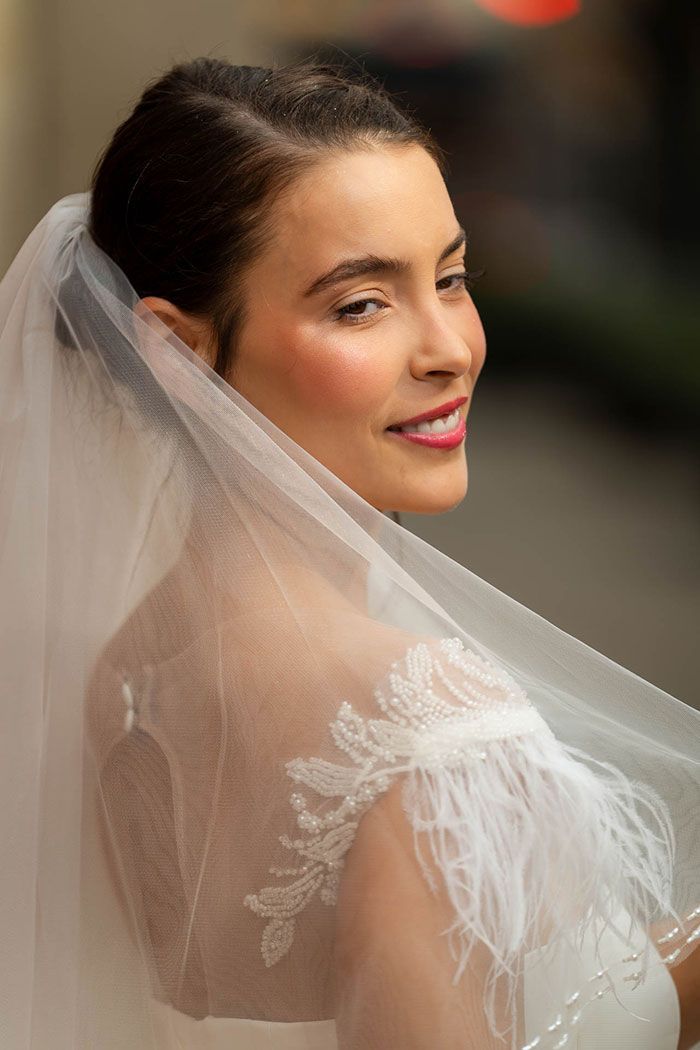 Novia sonriente, luciendo un velo y un vestido con encaje y plumas.
