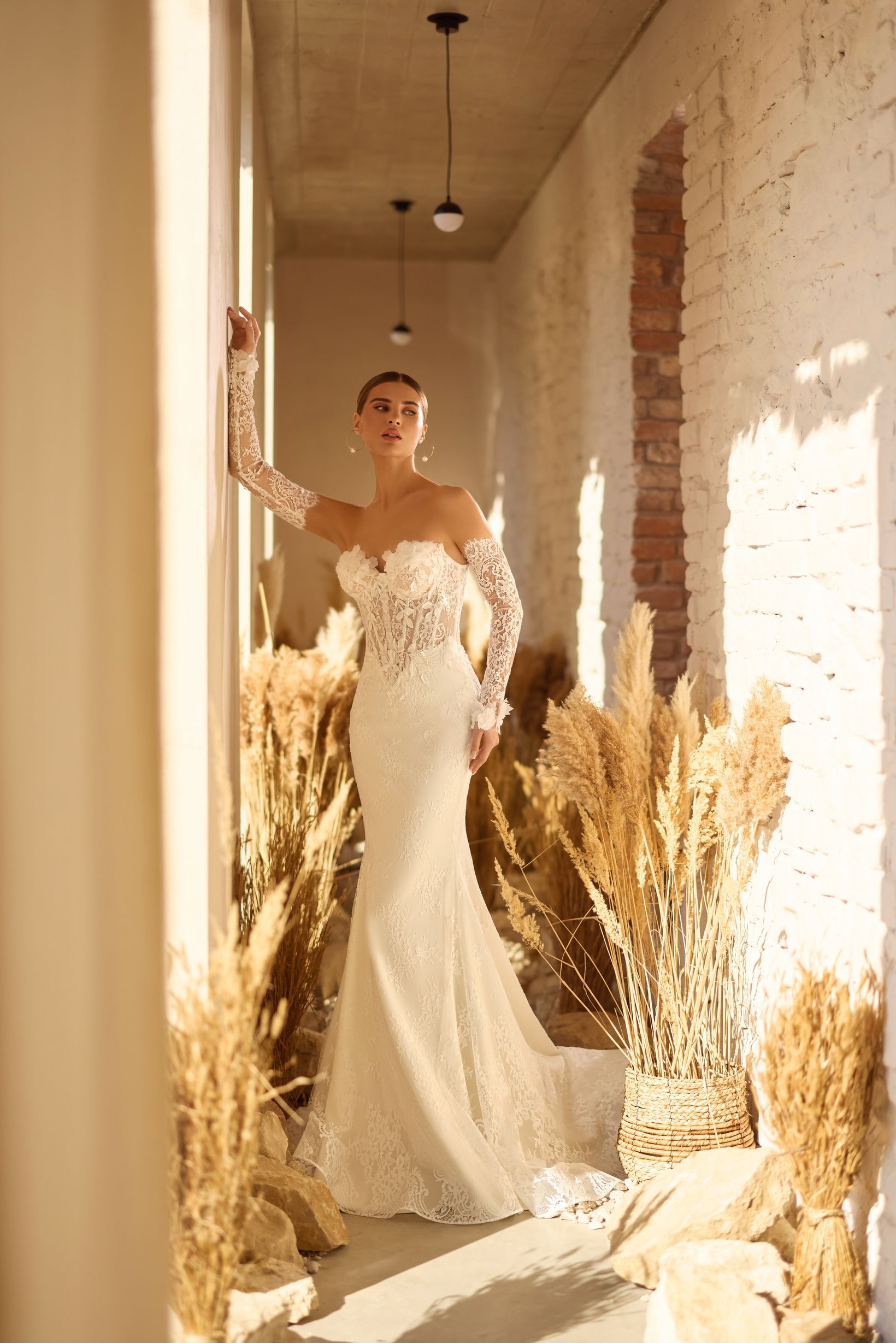 Mujer con vestido de novia de encaje blanco, apoyada en la pared. Plantas altas y secas al fondo.