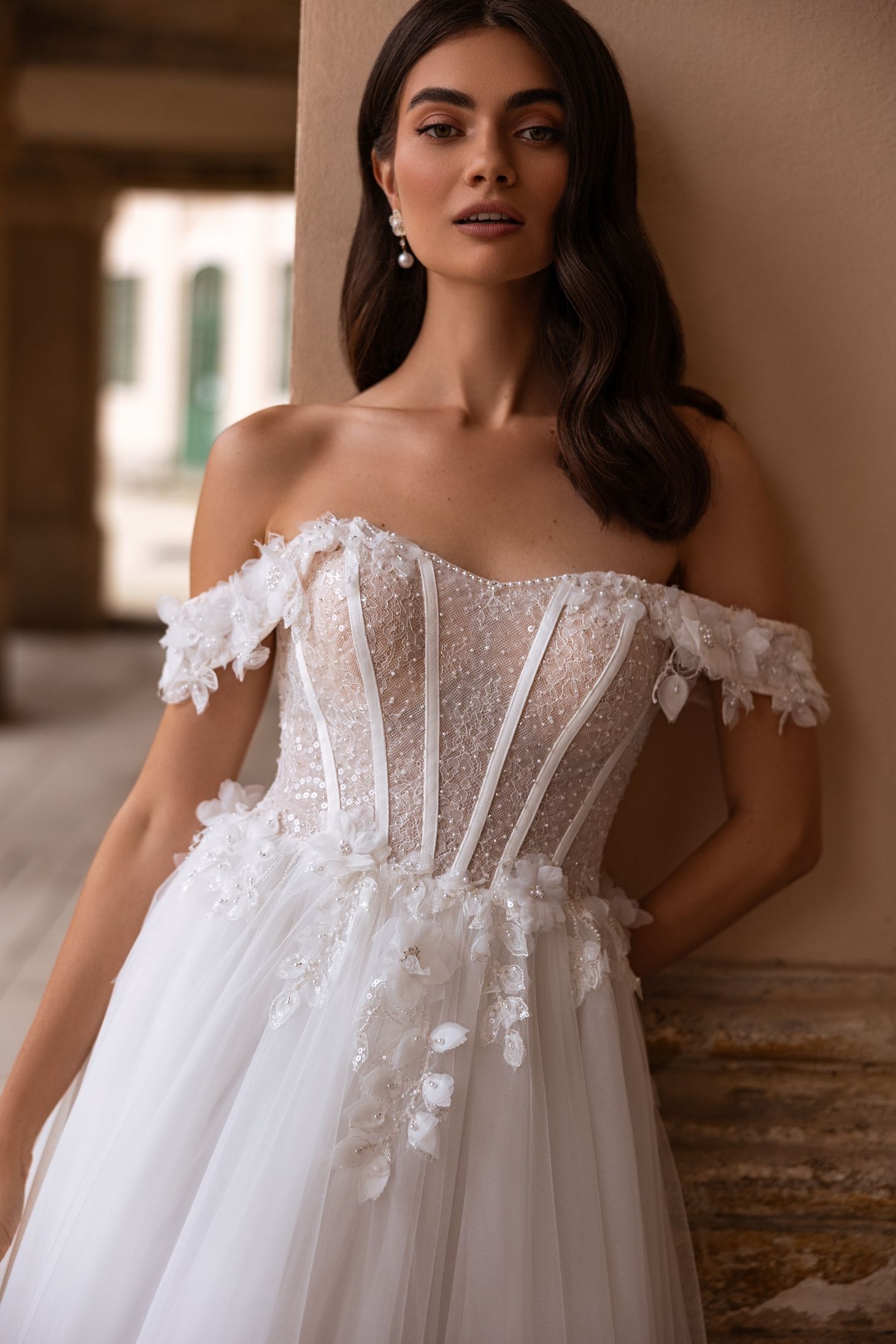 Mujer con un vestido de novia blanco con hombros descubiertos y apliques florales, de pie al aire libre cerca de una columna.