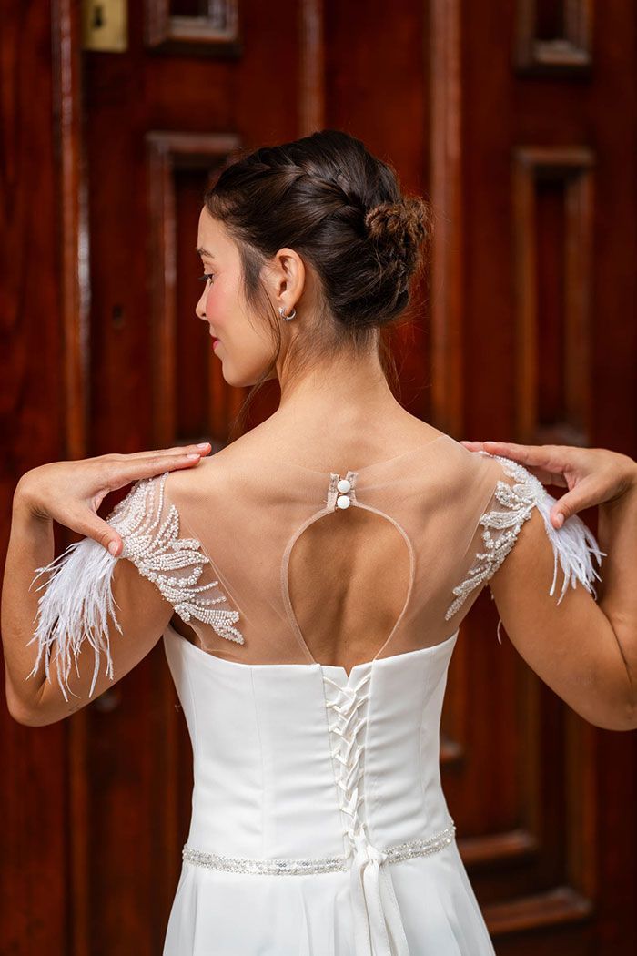 Novia con vestido blanco, brazos levantados, mostrando una espalda detallada con transparencias y detalles de plumas.