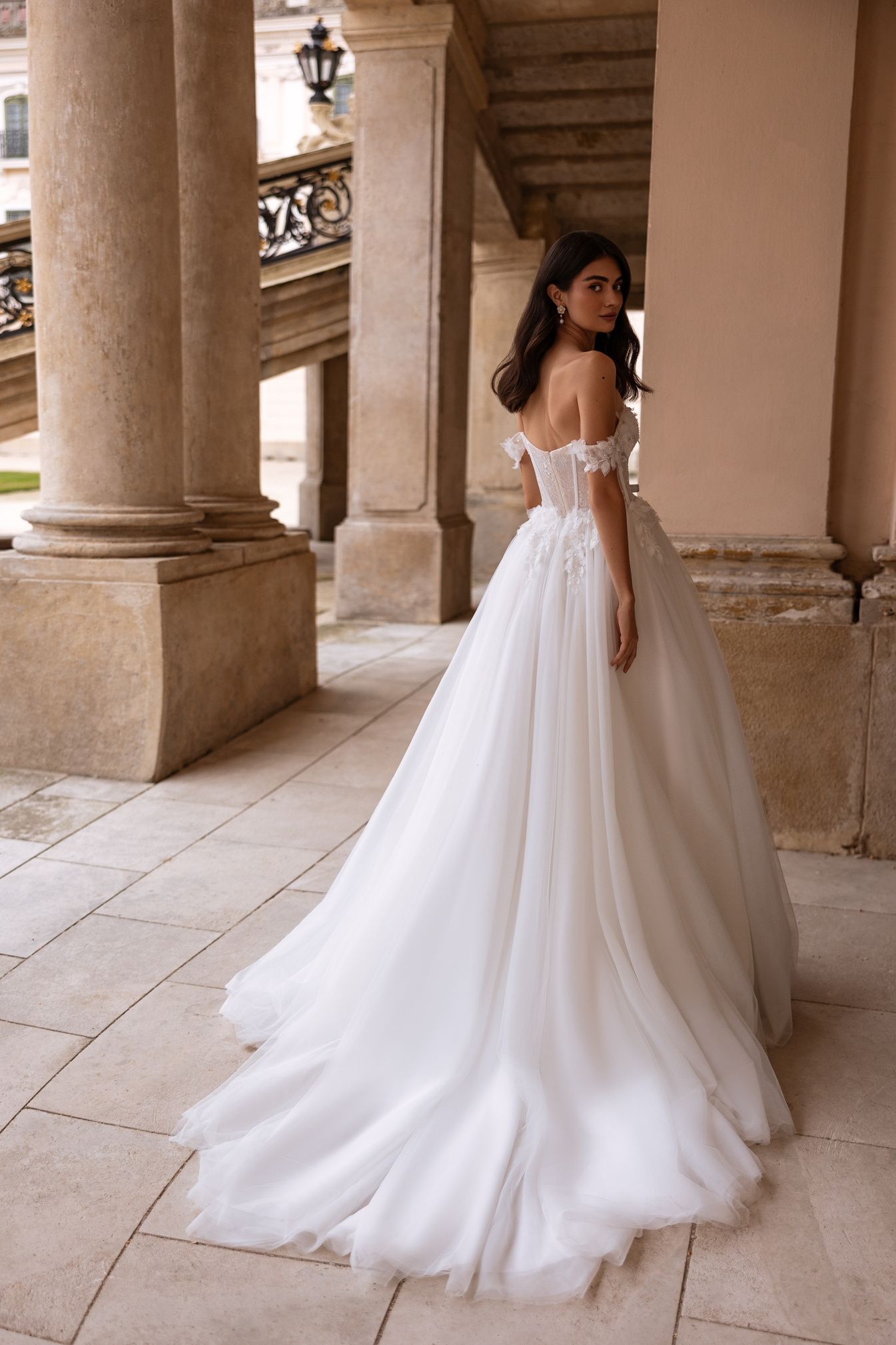Una mujer con un vestido de novia blanco con hombros descubiertos y una larga cola posa al aire libre cerca de columnas de piedra.