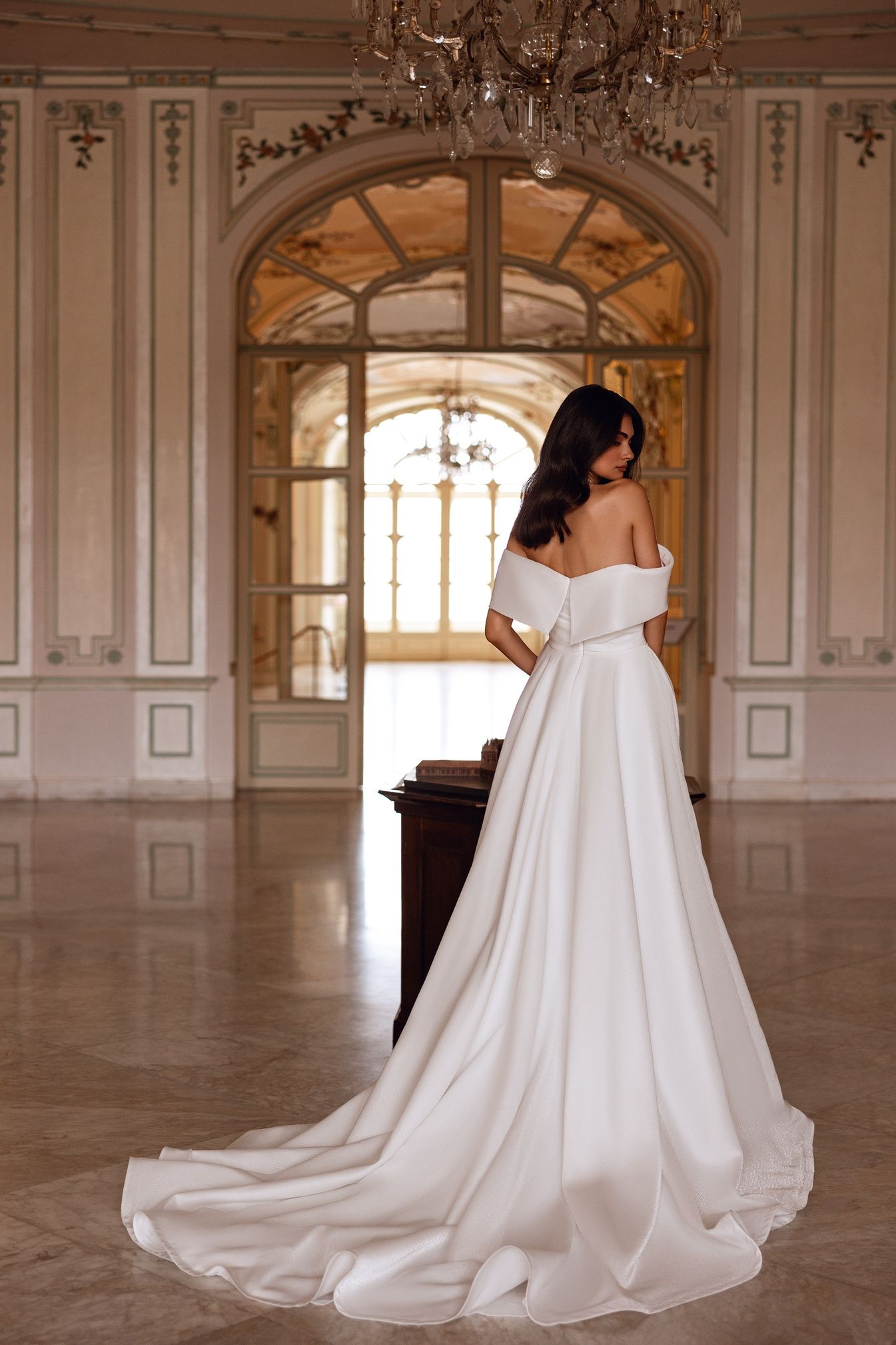 Una mujer con un vestido blanco con hombros descubiertos se encuentra en una gran habitación con grandes ventanas, mirando por encima del hombro.