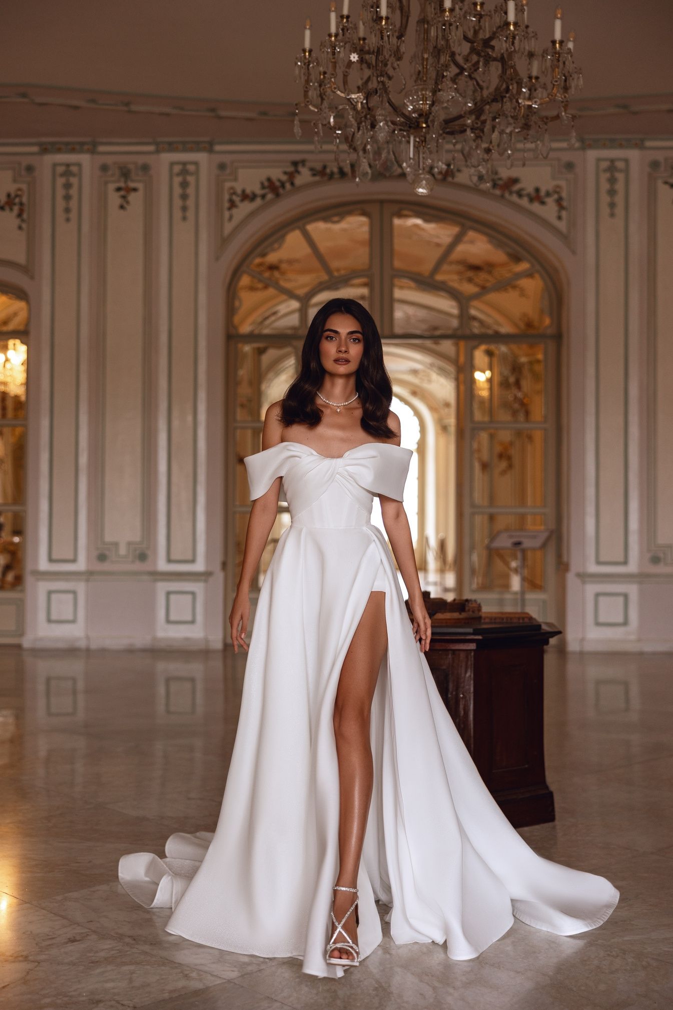 Mujer con un vestido blanco con hombros descubiertos y una abertura alta, de pie en un gran salón con pisos de mármol.