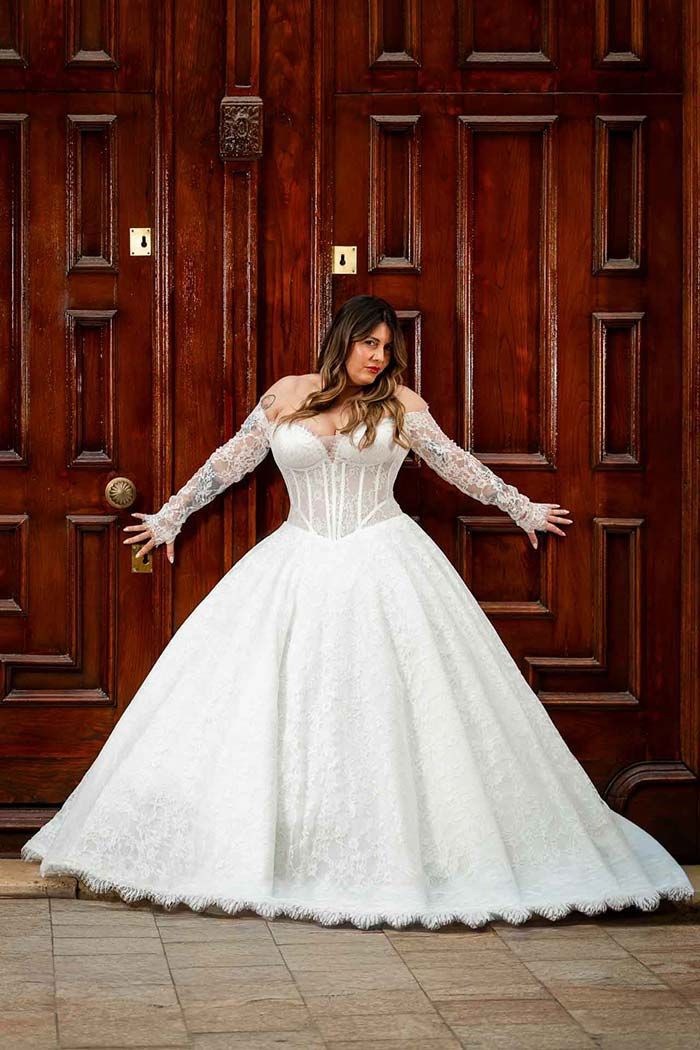 Una mujer con un vestido de encaje blanco se encuentra frente a unas grandes puertas de madera, con los brazos extendidos.