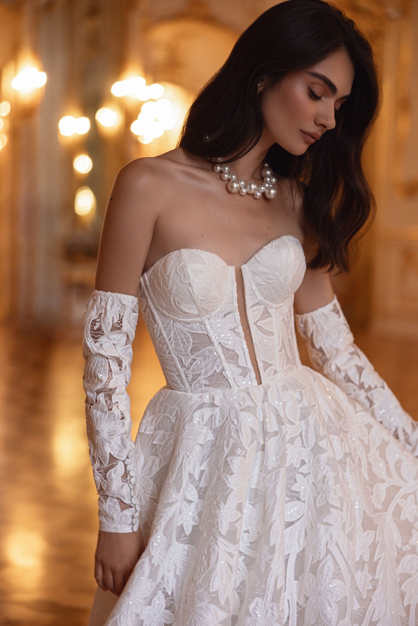 Mujer con un vestido de encaje blanco sin tirantes y guantes hasta el codo, luciendo un collar de perlas, en un salón de baile.