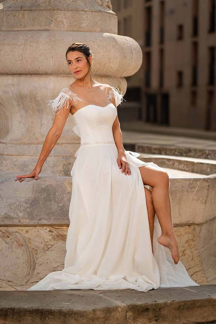 Mujer con vestido de novia blanco con abertura alta, sentada sobre estructura de piedra, al aire libre.