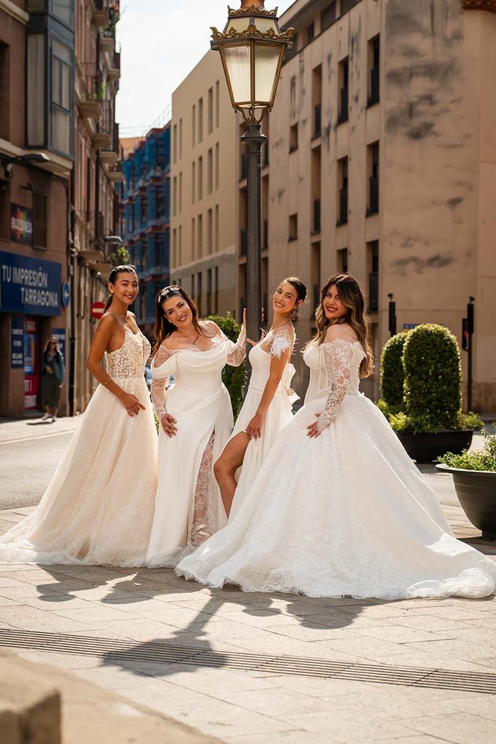 Cuatro personas con vestidos de novia, posando en una calle adoquinada cerca de una farola.