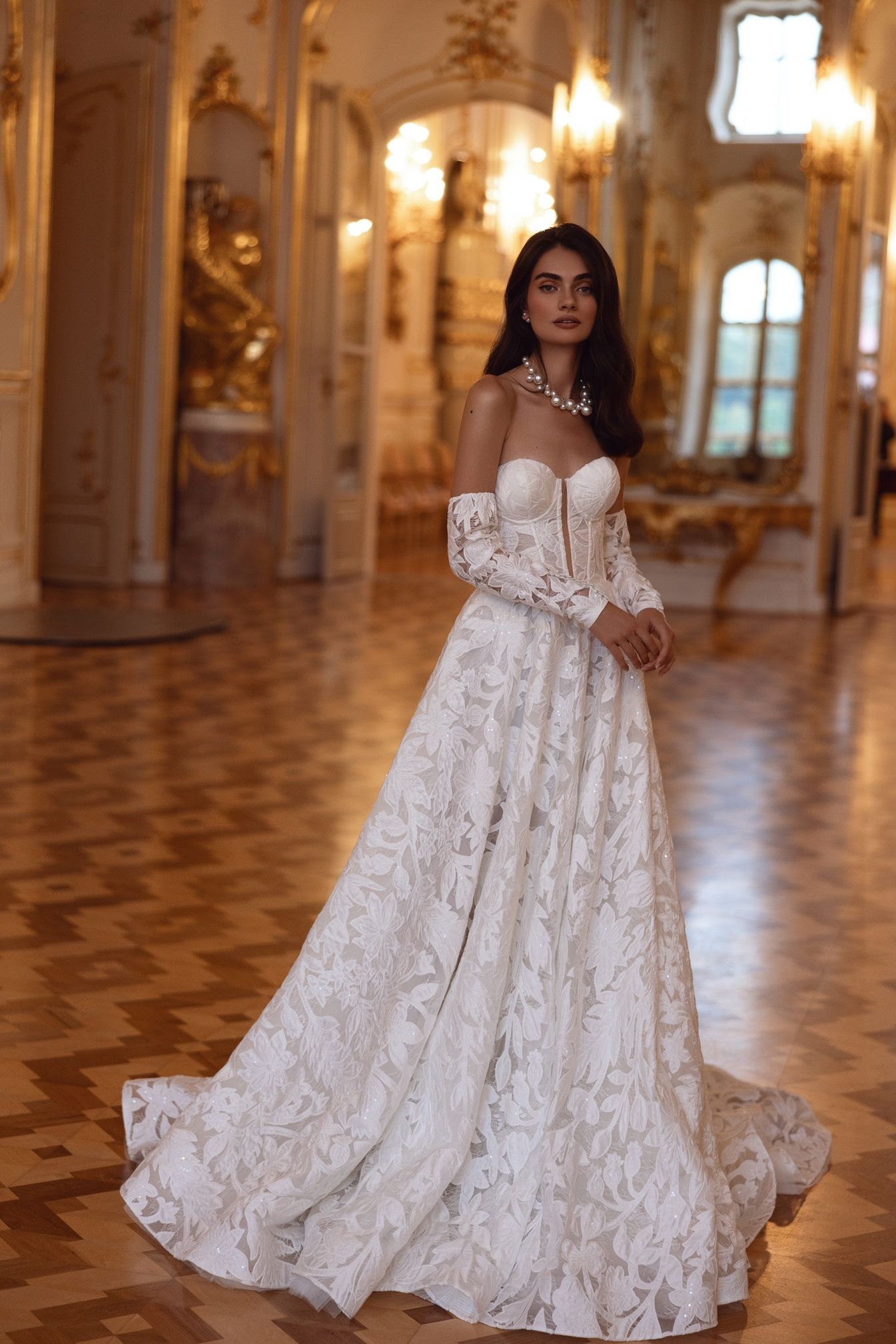 Mujer con un vestido de encaje blanco con mangas largas, de pie en un gran salón con una decoración ornamentada y un piso de madera.