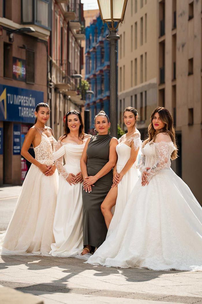 Cinco mujeres con vestidos de novia posan en una calle adoquinada. Una mujer con un vestido oscuro está de pie en el centro.