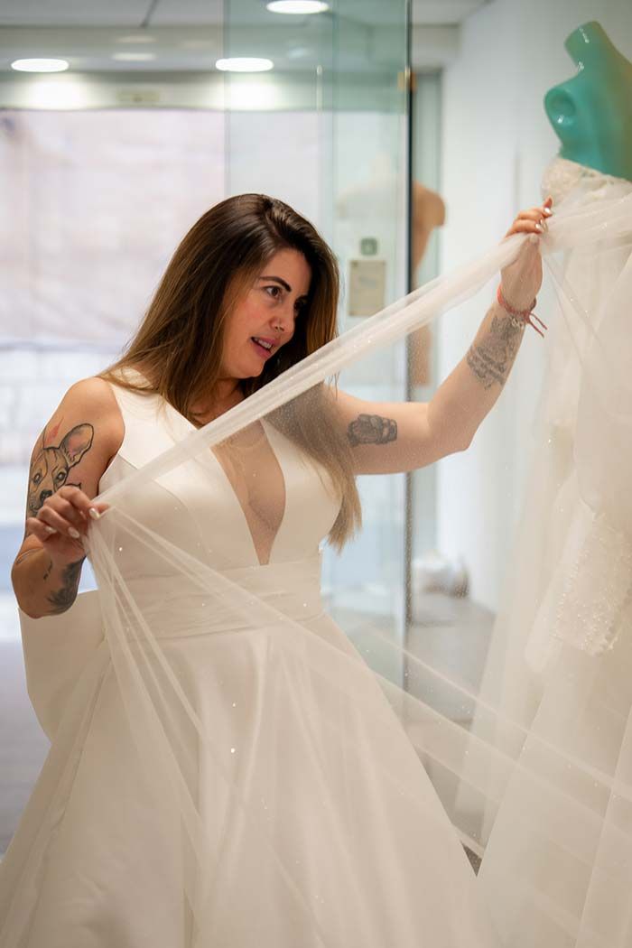 Mujer sosteniendo una tela transparente, admirando un vestido de novia blanco en una tienda de novias.
