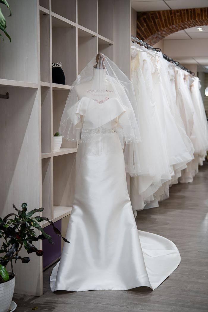 Vestido de novia sobre un maniquí con velo en una tienda de novias; otros vestidos cuelgan al fondo.