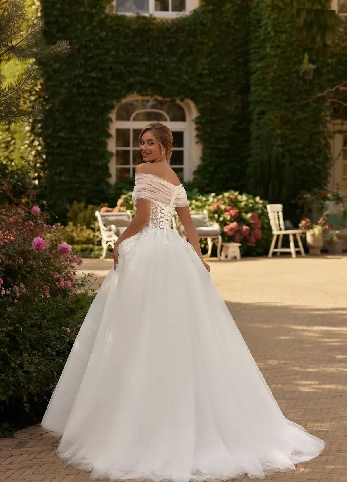 Una novia con un vestido blanco con hombros descubiertos posa frente a un edificio cubierto de enredaderas.
