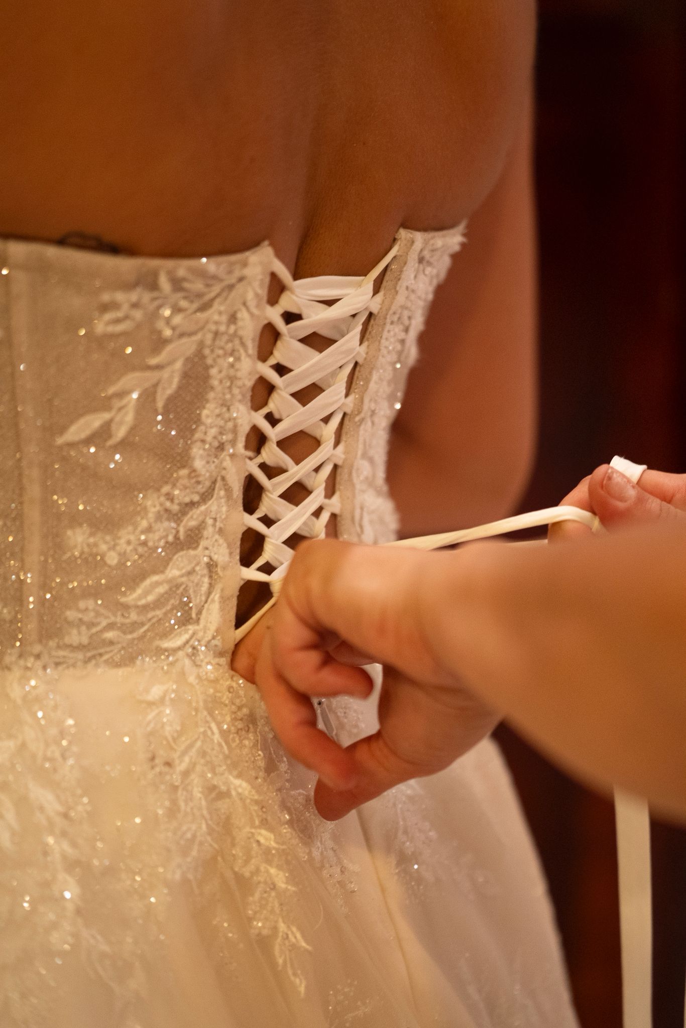 Espalda de una mujer con un vestido de novia blanco brillante, atado por una persona de piel clara.