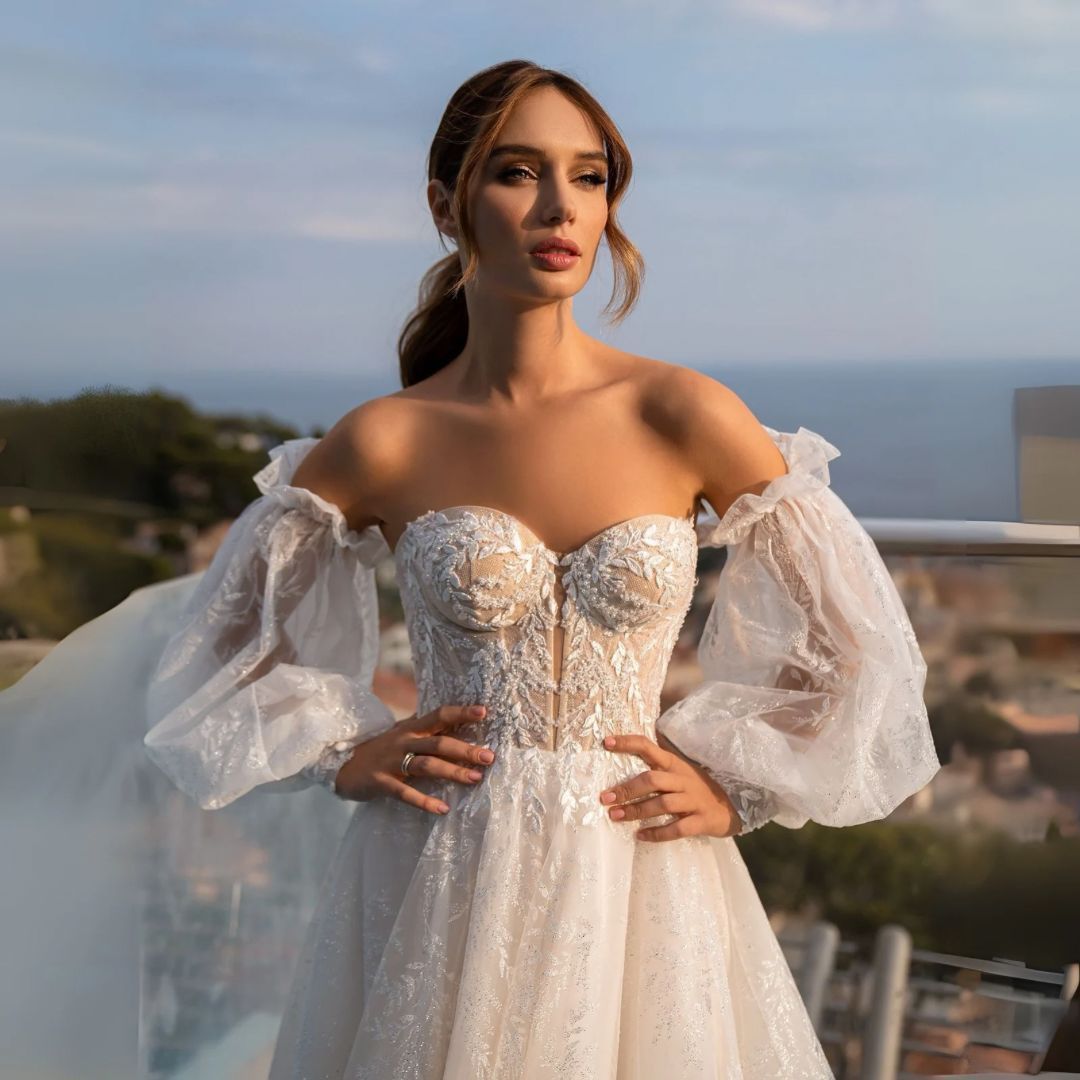 Mujer con vestido de novia con mangas de hombros descubiertos, al aire libre con el océano de fondo.