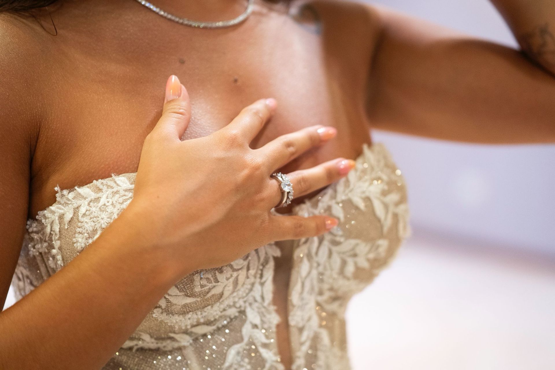 Mujer con vestido de novia sin tirantes, mano en el pecho mostrando el anillo de compromiso.