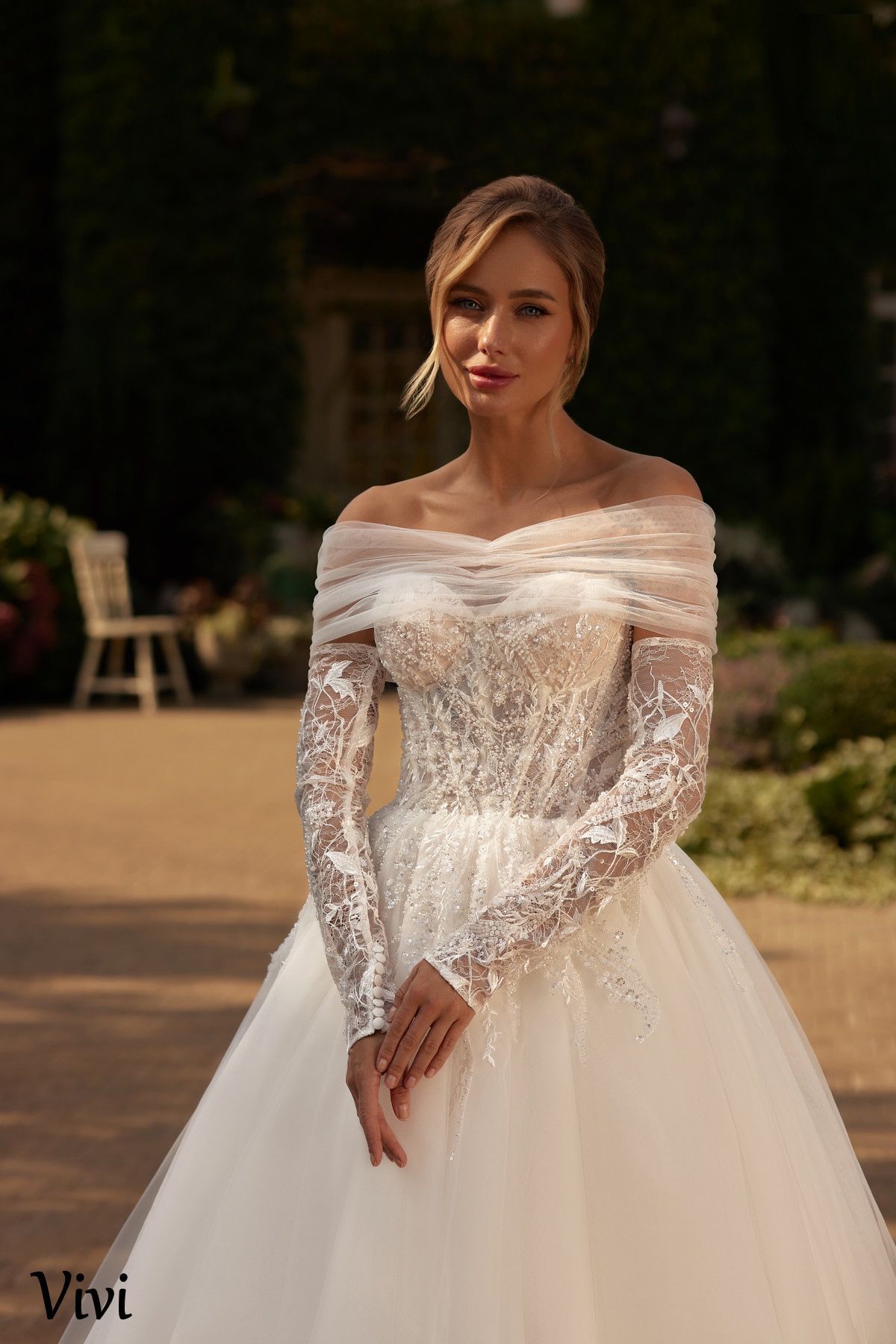 Mujer con un vestido de novia blanco de hombros descubiertos, sonriendo. Un espacio al aire libre con vegetación y una silla.