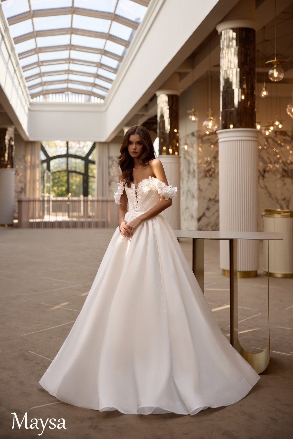 Una mujer con un vestido de novia blanco con hombros descubiertos posa en un gran salón con columnas de mármol y un tragaluz.