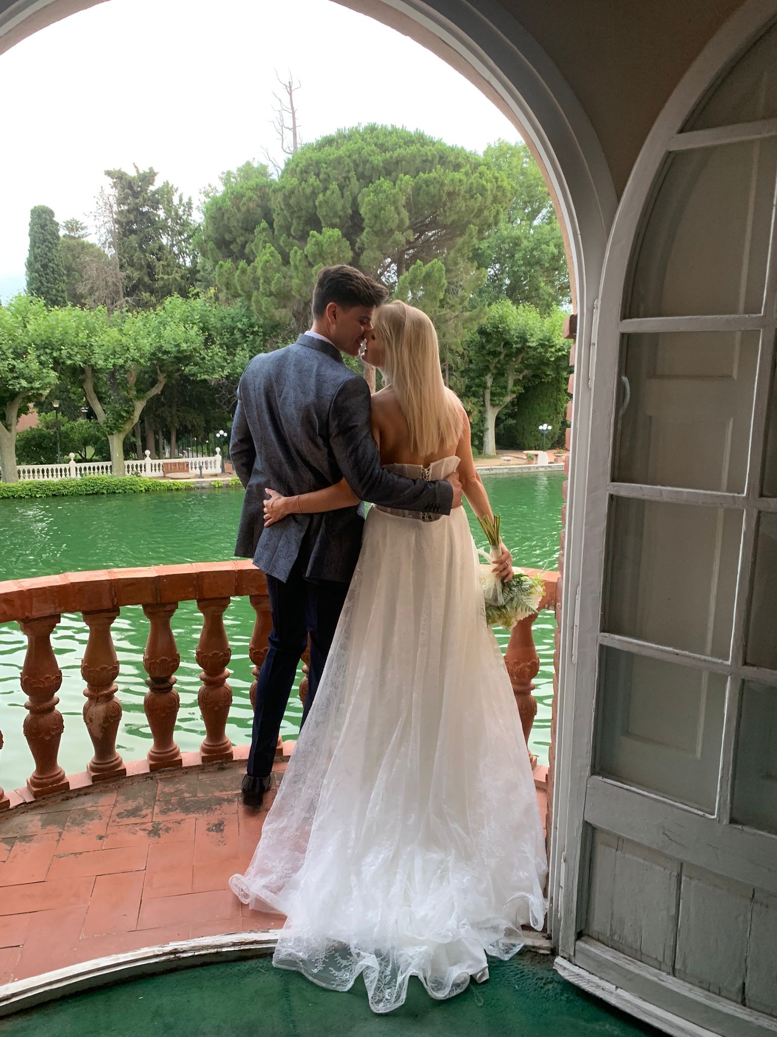 Pareja besándose en un balcón con vistas a un lago, la mujer con un vestido de novia, el hombre con traje.