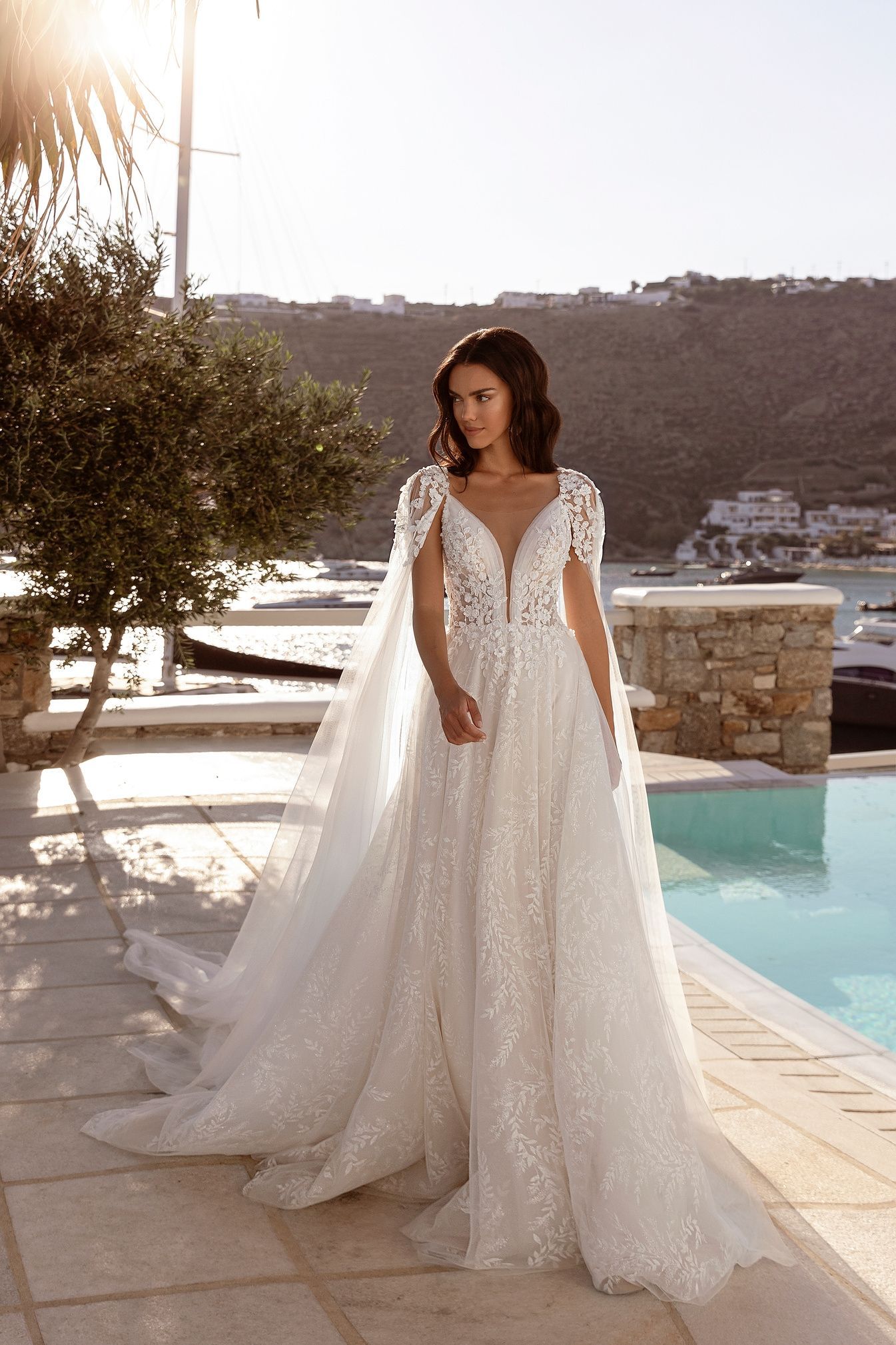 Mujer con vestido de novia blanco con capa, al aire libre cerca de una piscina y agua con sol.