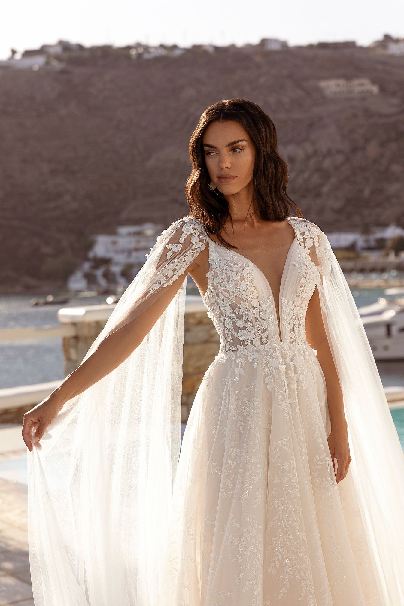 Mujer con vestido de novia y capa, posando al aire libre cerca de una piscina y agua.