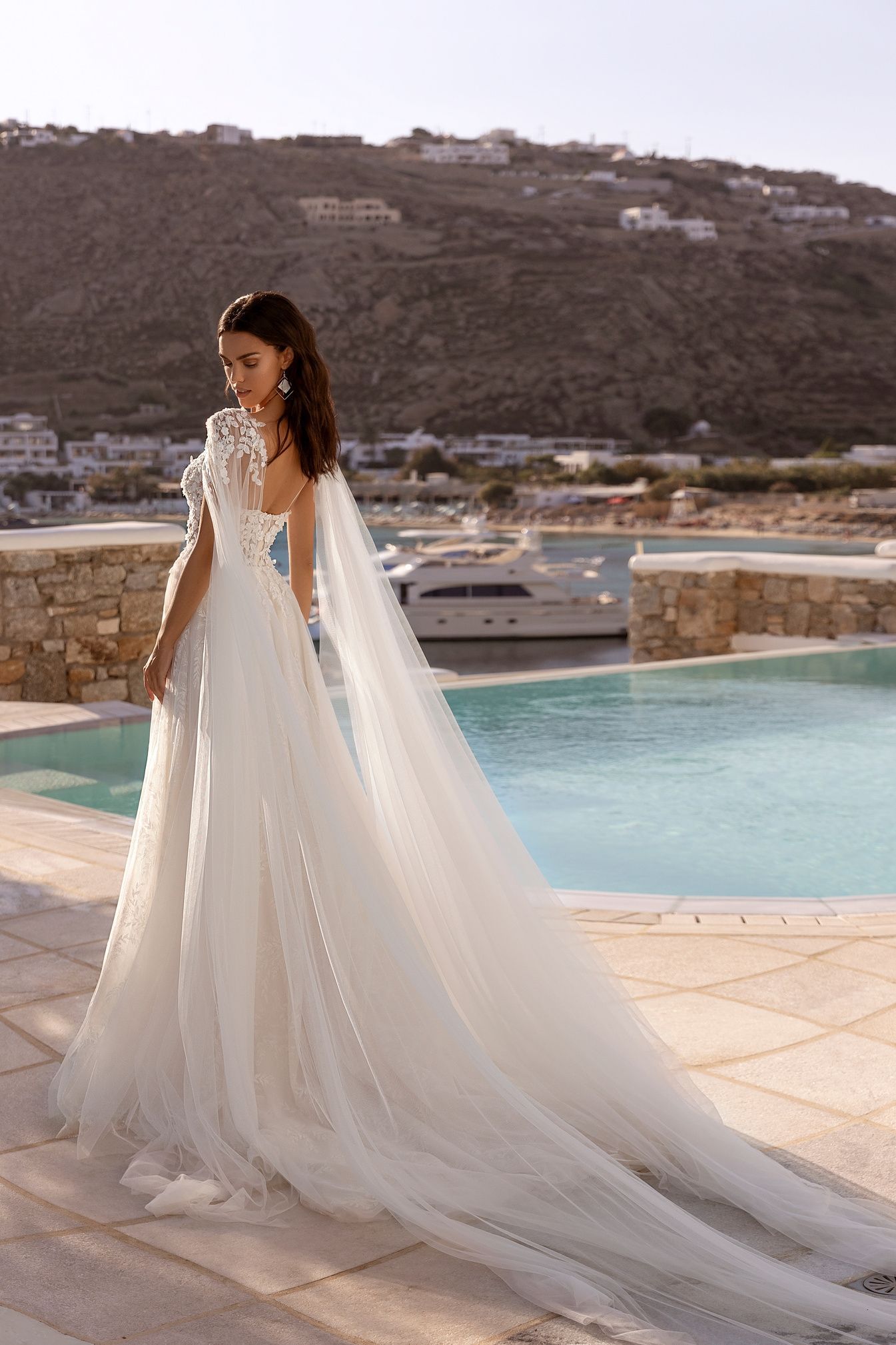 Mujer con un vestido de novia blanco con una larga cola, de pie cerca de una piscina con un fondo escénico.