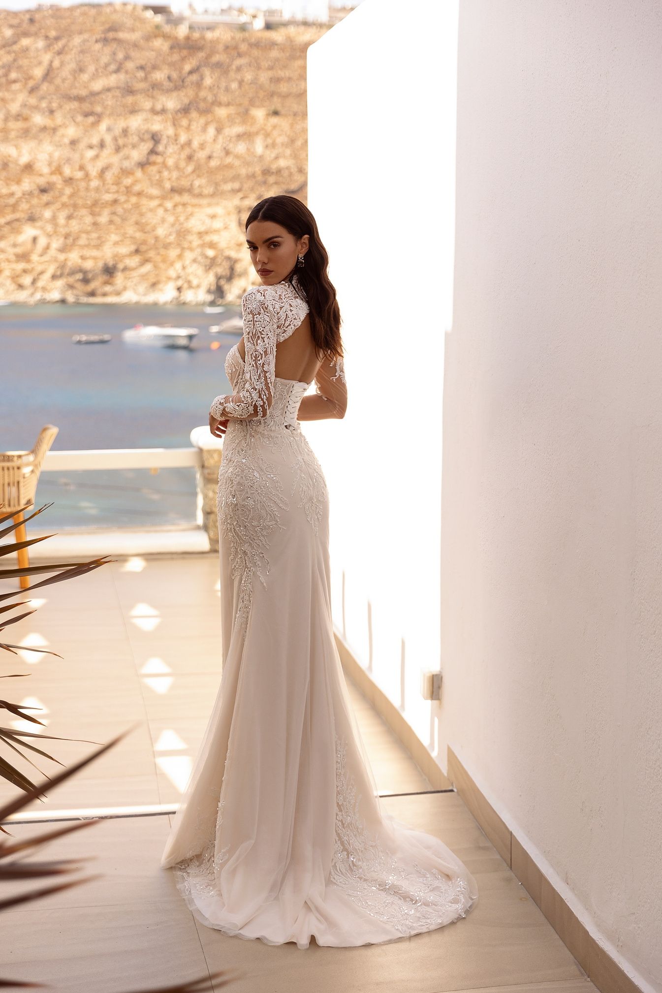 Mujer con un vestido de novia blanco, de pie en un balcón, con vista al mar y a la montaña.