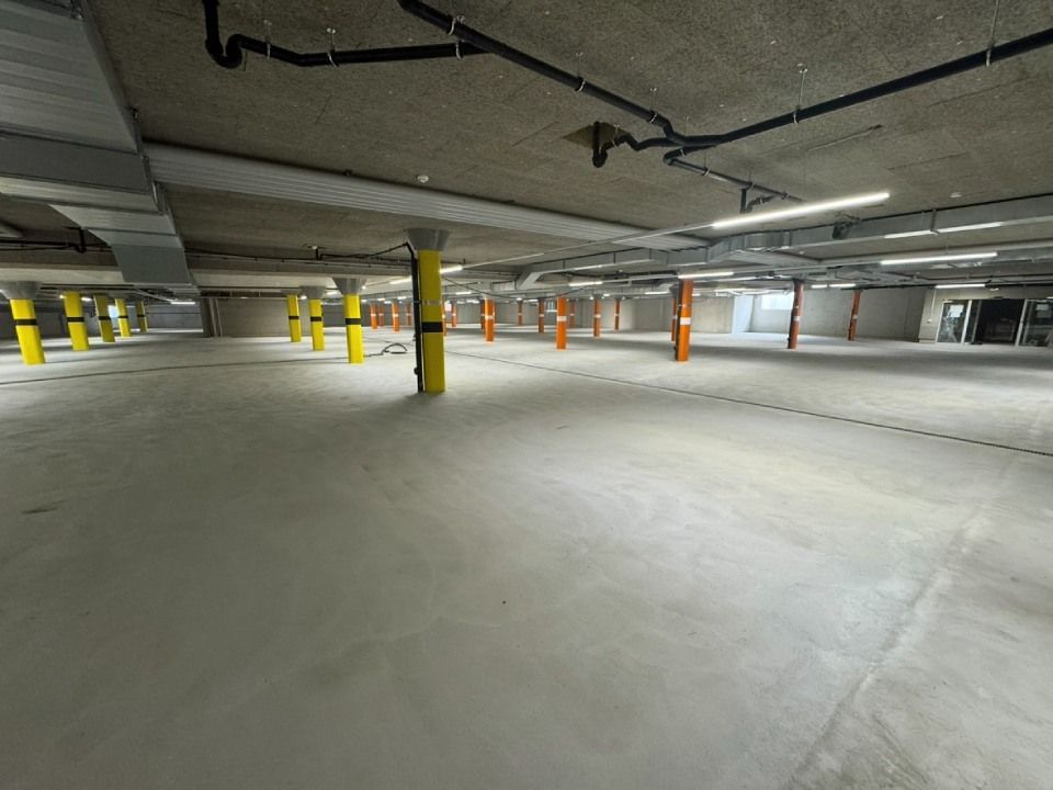 Leere, geräumige Tiefgarage mit Betonpfeilern und -decke, darüberliegenden Rohren und Leuchtstoffröhren.