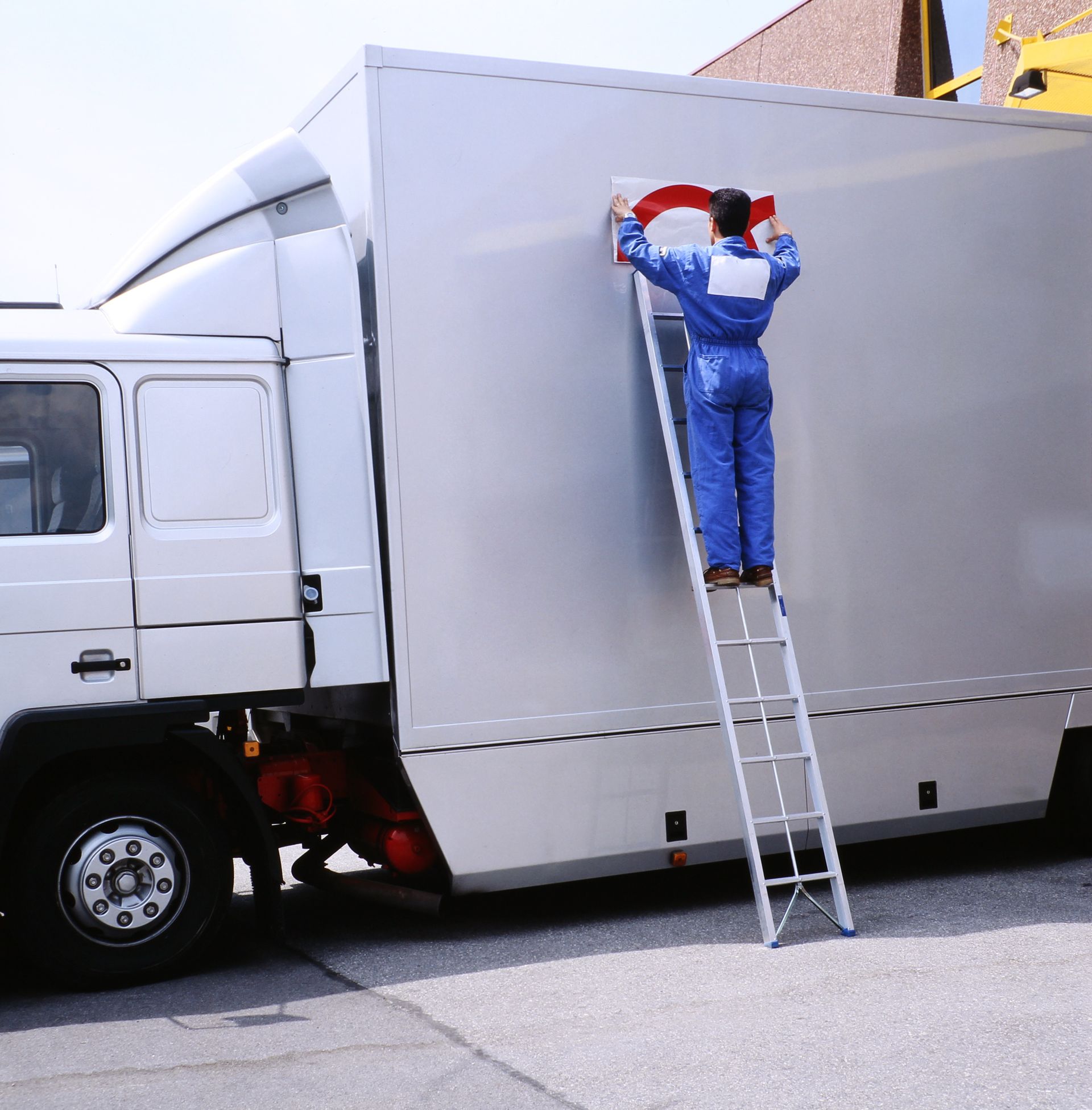 Un ouvrier, juché sur une échelle, applique de la peinture rouge sur la remorque d'un grand camion blanc.