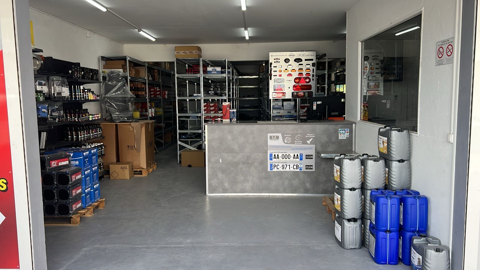 Intérieur d'une zone de stockage d'entrepôt avec des étagères métalliques, des cartons empilés et des conteneurs bleus près de l'entrée.