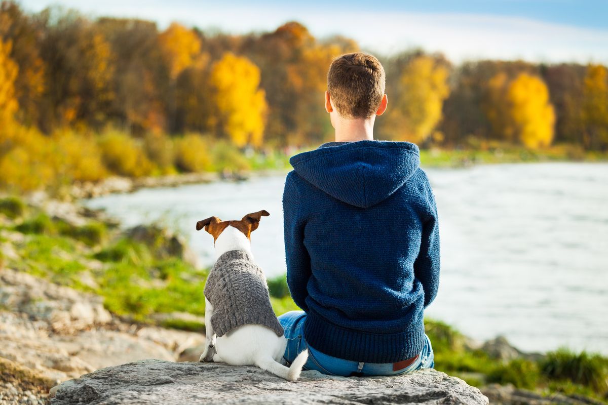 Una persona y un perro pequeño sentados en una roca junto a un lago, frente a árboles otoñales con follaje amarillo.