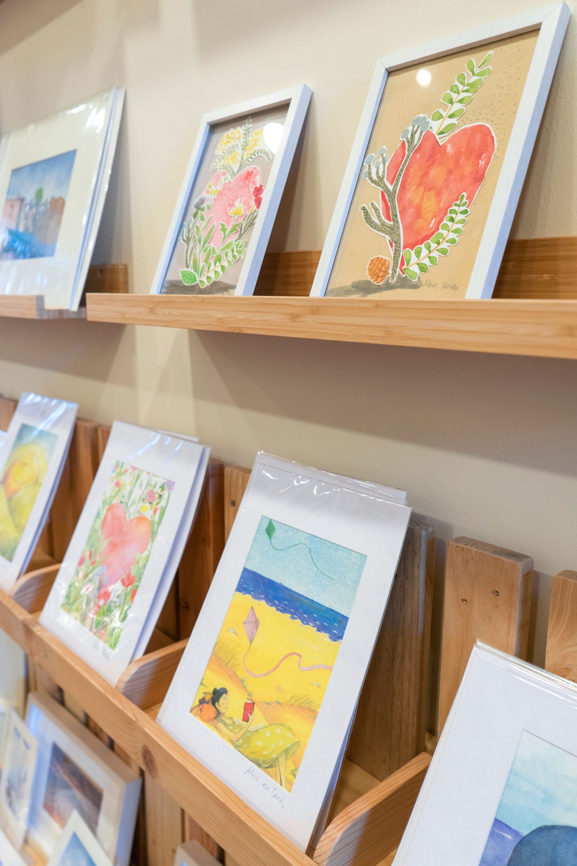 Obras de arte en marcos blancos expuestas en estantes de madera, pinturas de acuarela con corazones y paisajes.