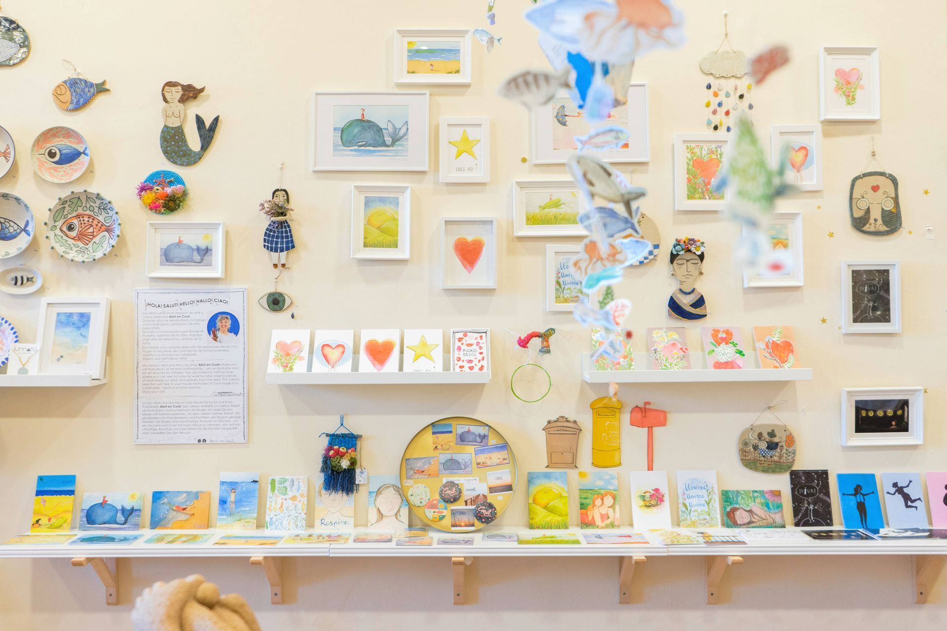 Una exhibición de arte colorida con dibujos enmarcados, cerámicas y tarjetas en estantes contra una pared de color crema.