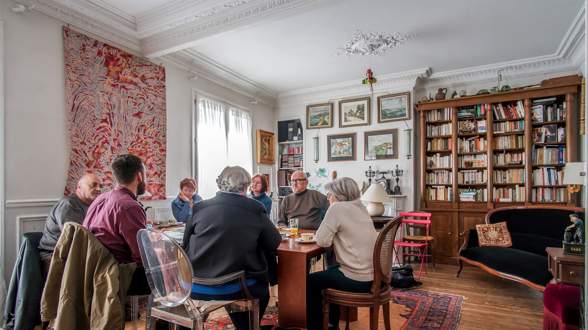 Un groupe de personnes est assis autour d'une table, dans une pièce. On aperçoit une bibliothèque, des tableaux au mur et une tapisserie.